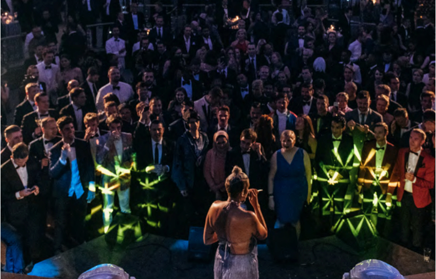 A singer performs for well-dressed audience under chandeliers.